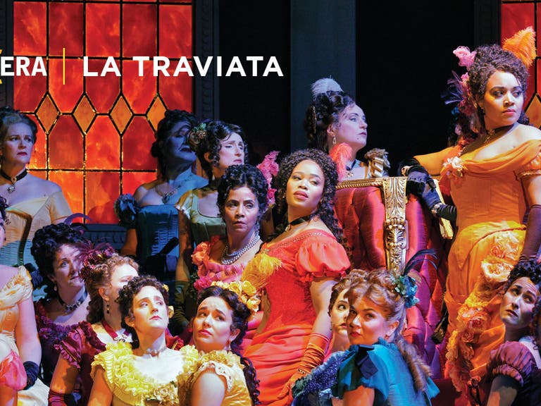 Group of women in colorful dresses looking to one side. Orange stained glass in the background. Text reads LA Opera La Traviata.