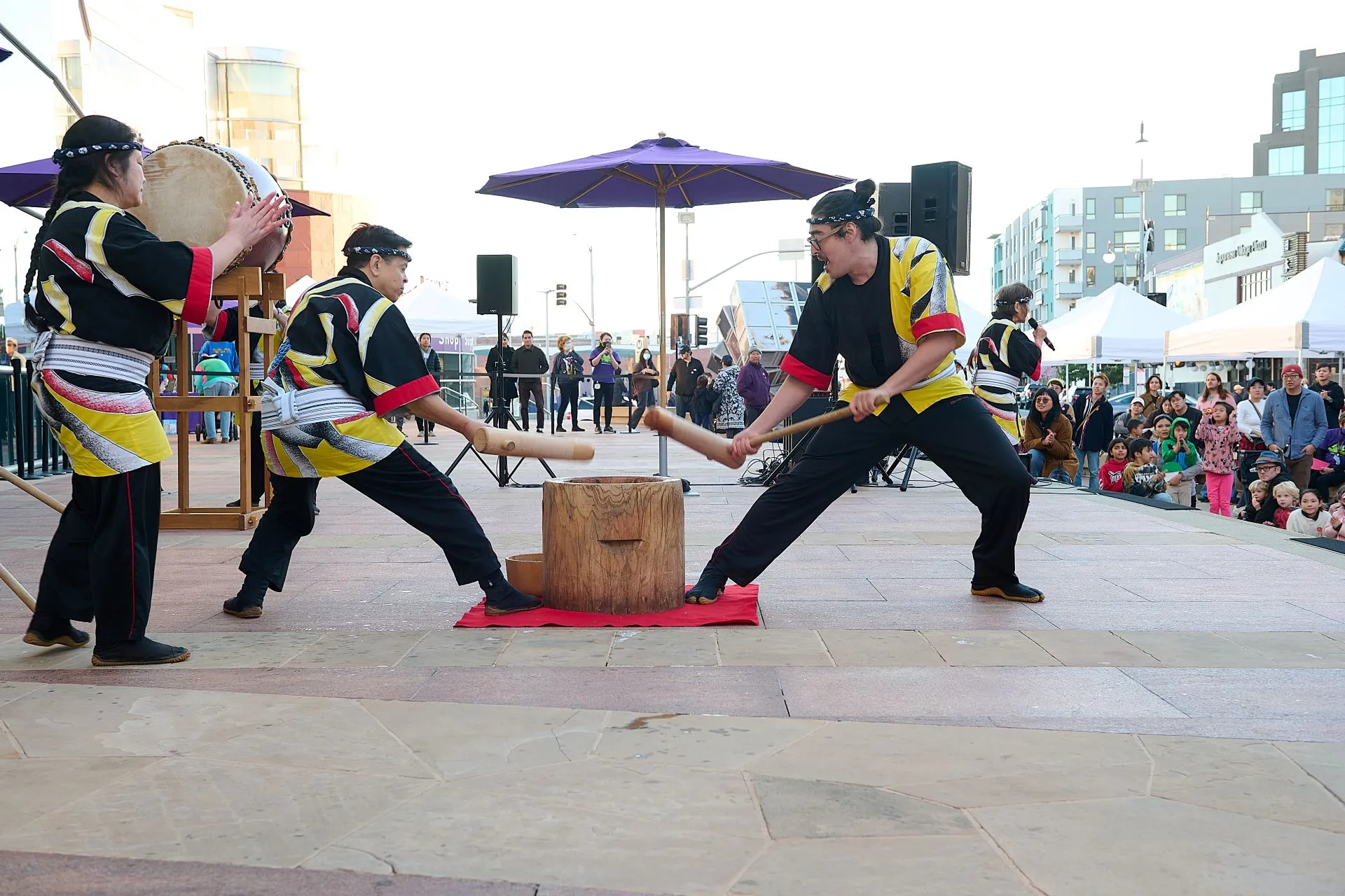 Two Taiko (Japanese drum) performers pounding a large wooden container of rice with mallets in a mochitsuki (mochi making) demonstration.