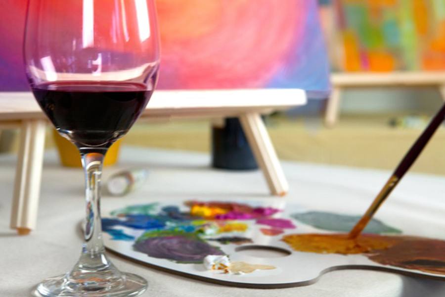 A wine glass and a pallet with paint sit in front of a painting on an easel