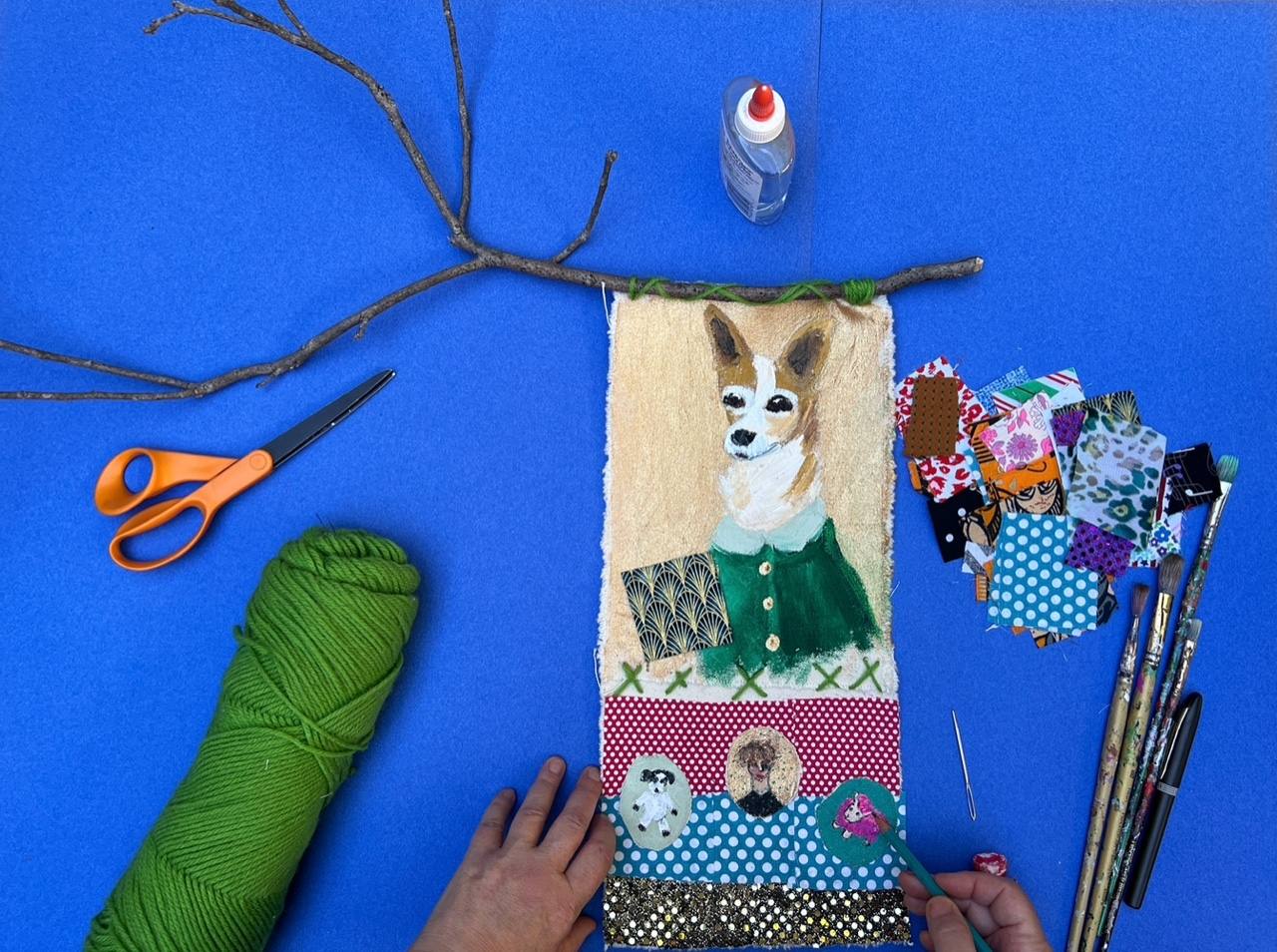 Image of hands creating a portrait of a dog using fabrics, thread, and paint.