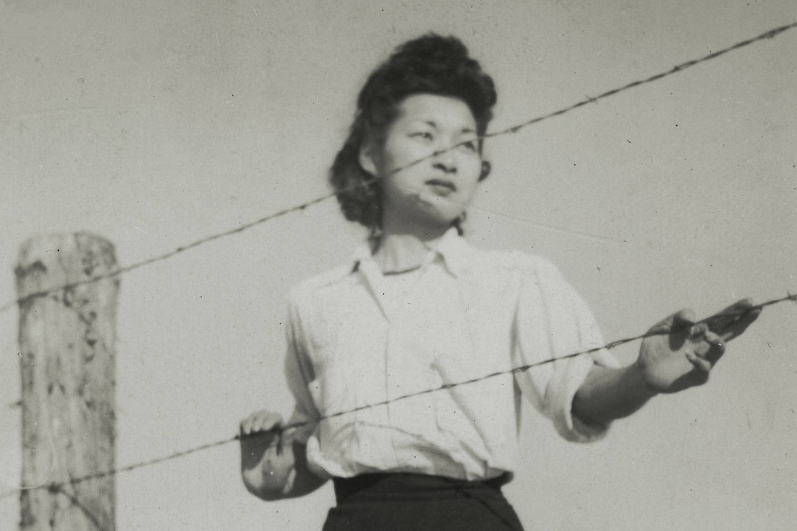 a young woman standing by the barbed wire fence of the Manzanar concentration camp. Anonymous. Gift of Myrtle Joyce Barley Ward, JANM. 2003.12.14.