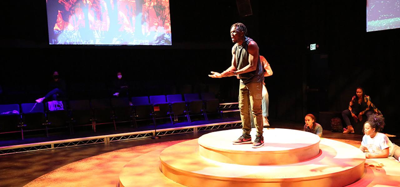 Man stands in the center of a circular stage, under a spotlight, with his hands outstretched.