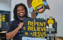A man poses with a sign that says "Repent and Believe in Jesus" that has been altered into a piece of artwork featuring a laughing Jesus emoji