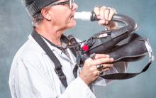 an artist named phog masheen, wearing many rings and a hat, wearing a white overshirt in front of a hazy blue background has lips pursed against an unusual instrument made of pipes.