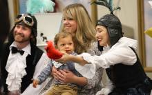 Two performers in costume with feathers and aviator goggles, sitting with a mother and child. The adults are smiling, the baby is sucking his thumb and looking at one of the performers who is showing the baby a red plush bird. 