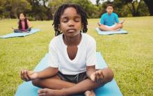 child sits in a meditative pose on a light blue yoga mat in a grassy field (this is not the venue)